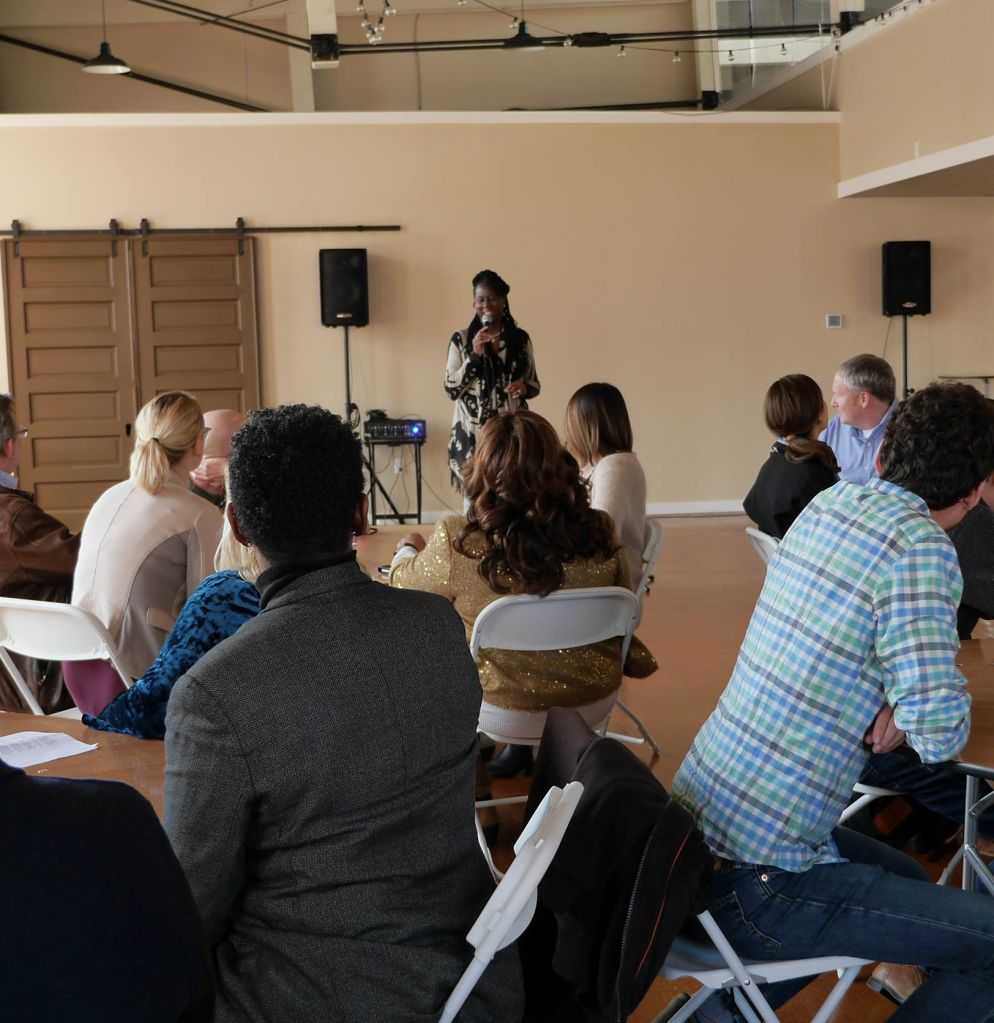 A facilitator stands at the front of a room of people sitting at circular tables